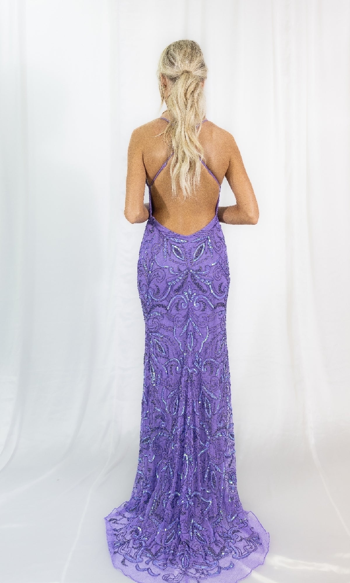 A blonde-haired woman in a ponytail models the Long Sequin-Print Prom Dress with a strappy open back, floor-length silhouette, and sparkling details. She stands against a white backdrop, facing away from the camera.